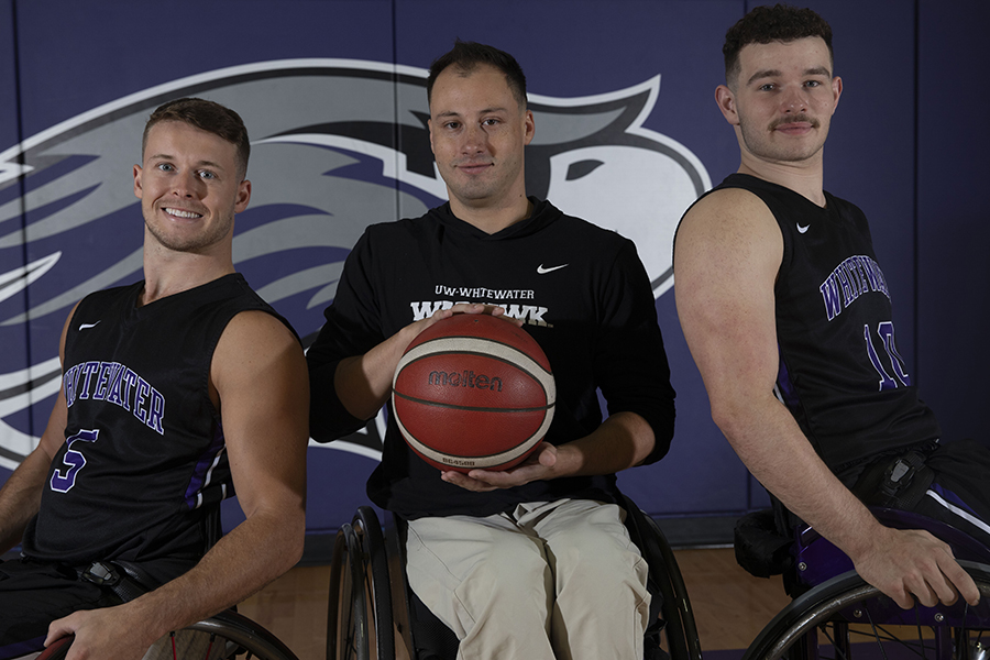 Three Warhawks pose in front of a Warhawk wall and hold a basketball.