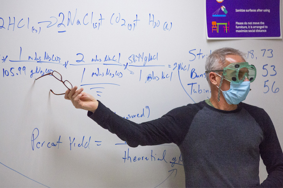 A faculty member, wearing safety goggles and a face mask, points at a formula on a whiteboard.