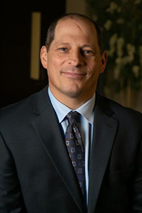 Andrae Marak sits in front of a dark office background wearing a suit and tie.