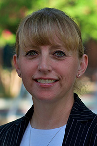 Kathryn Zuckweiler stands outside in front of green trees wearing a black suit coat over a white shirt.
