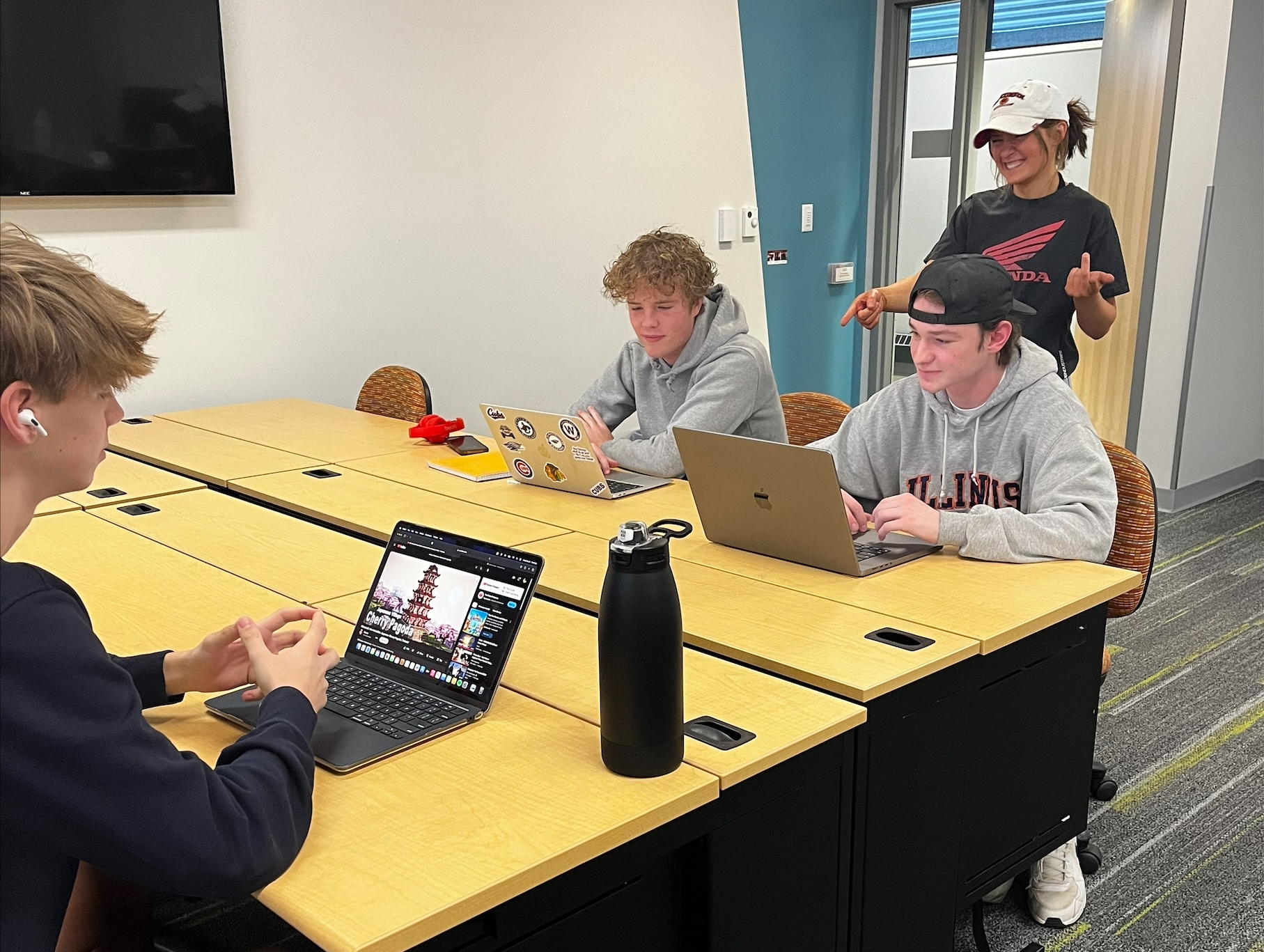 Students sitting at desks working on laptops with another student standing behind two of them.