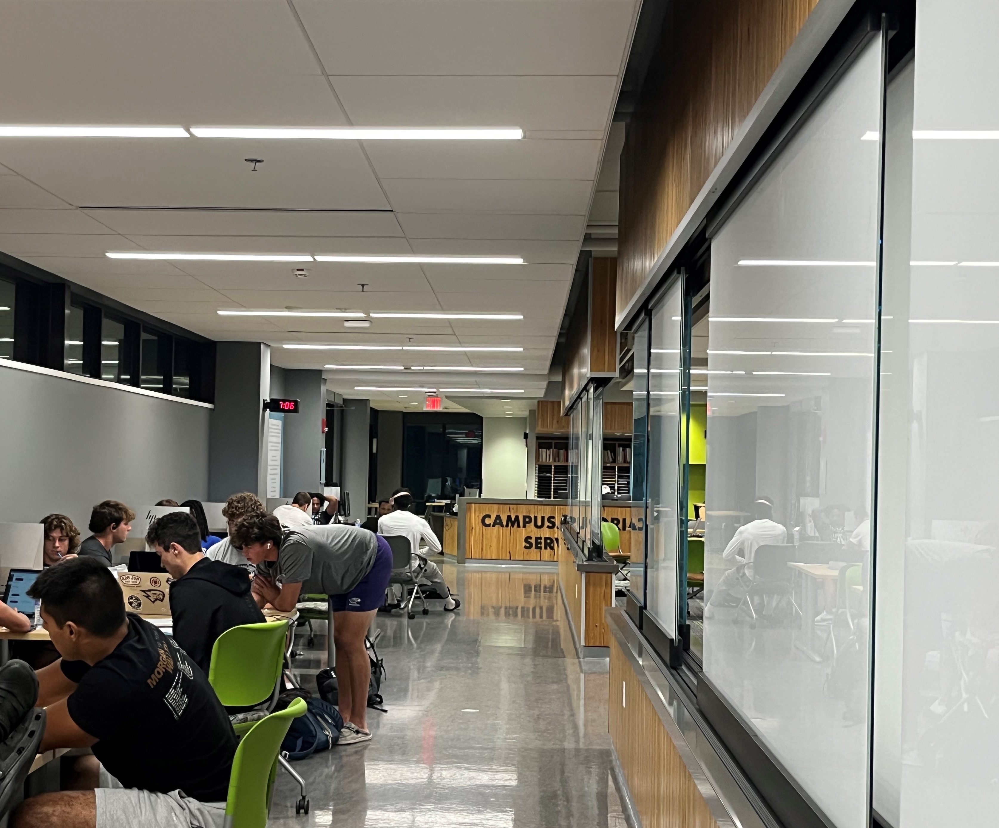 Students sitting at tables and doing work on their laptops in a wide hallway.