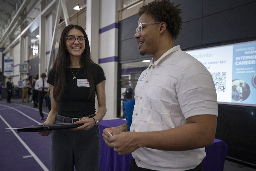 Two people wearing business casual attire chat and smile while holding folders.