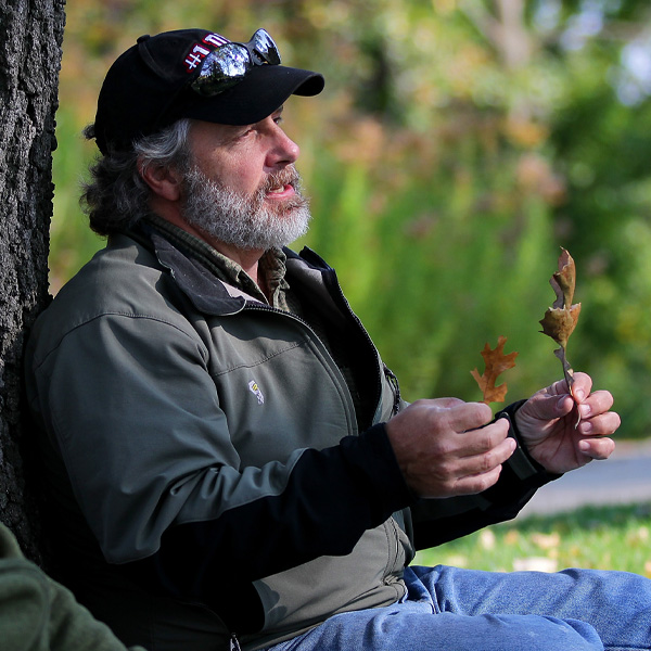 Tony Gulig sits with his back against a tree holding a leaf in each hand.