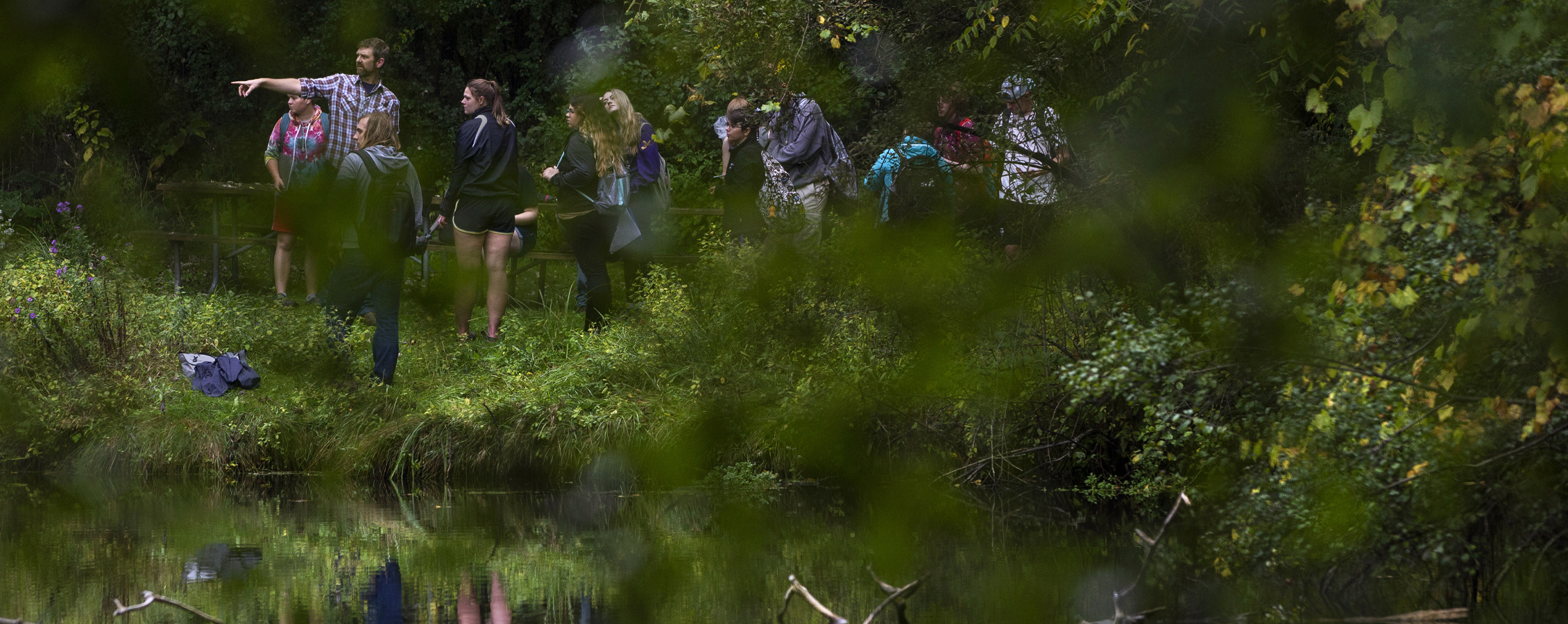 Students and a faculty member walk through a forest with a small body of water.