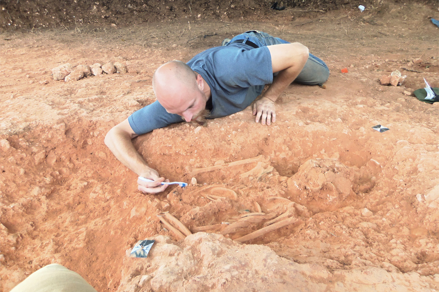 A student lays in the dirt over a skeleton that he's uncovering with a toothbrush.
