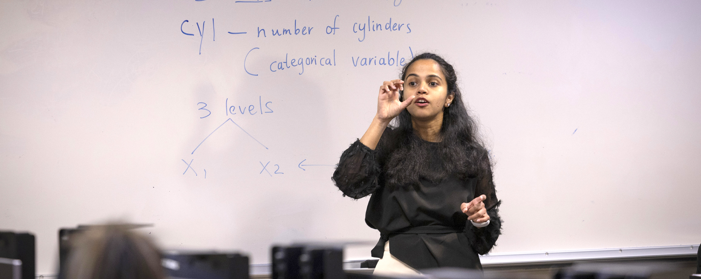 An instructor speaks in front of a white board with equations written on it.