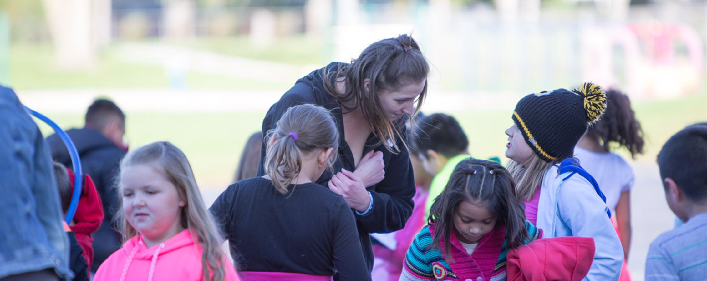 A school psychology student stands amongst a group of children and bends down to listen as a child speaks to them.