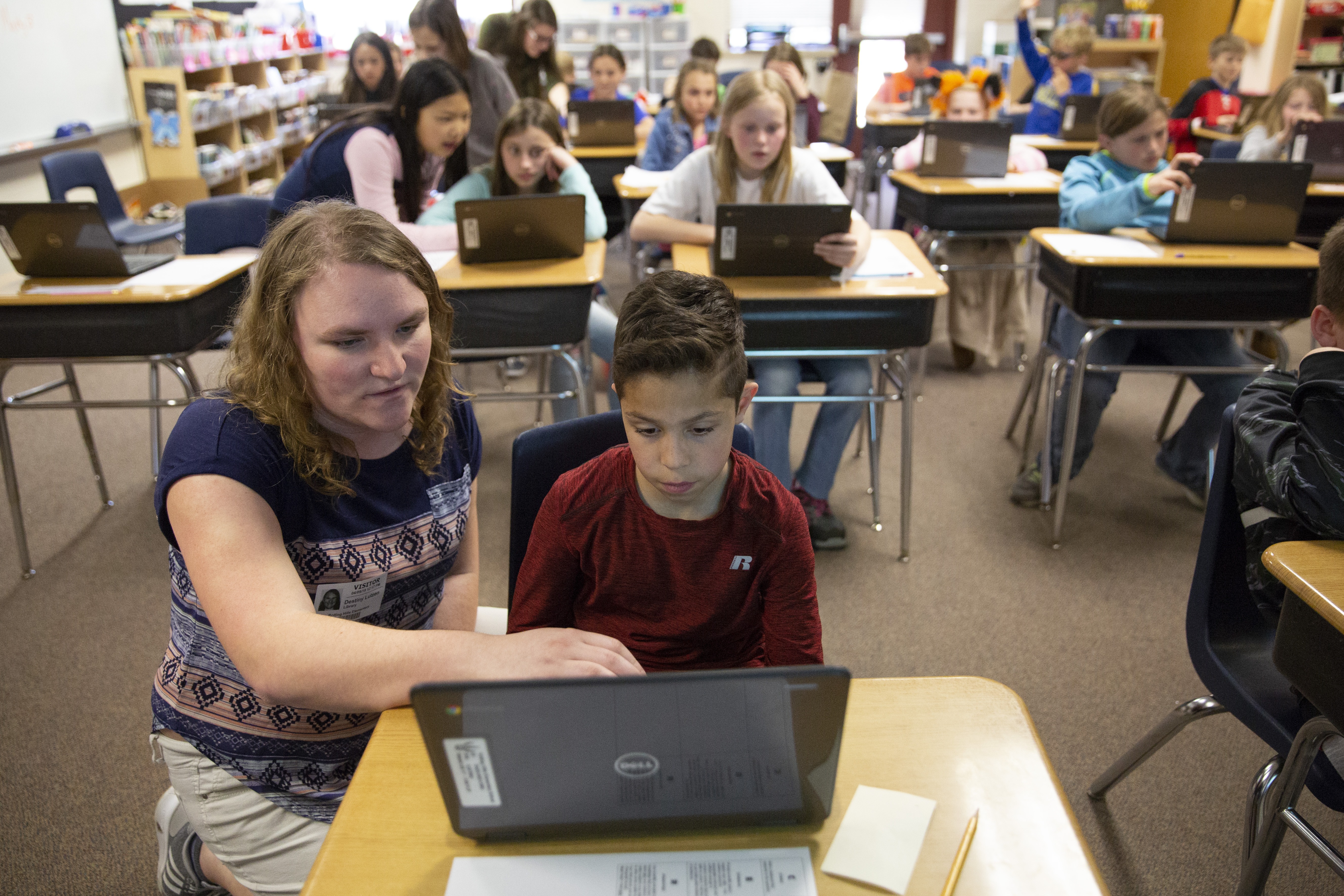 A student teacher kneels beside a young student sitting at a desk as they both look at a laptop.