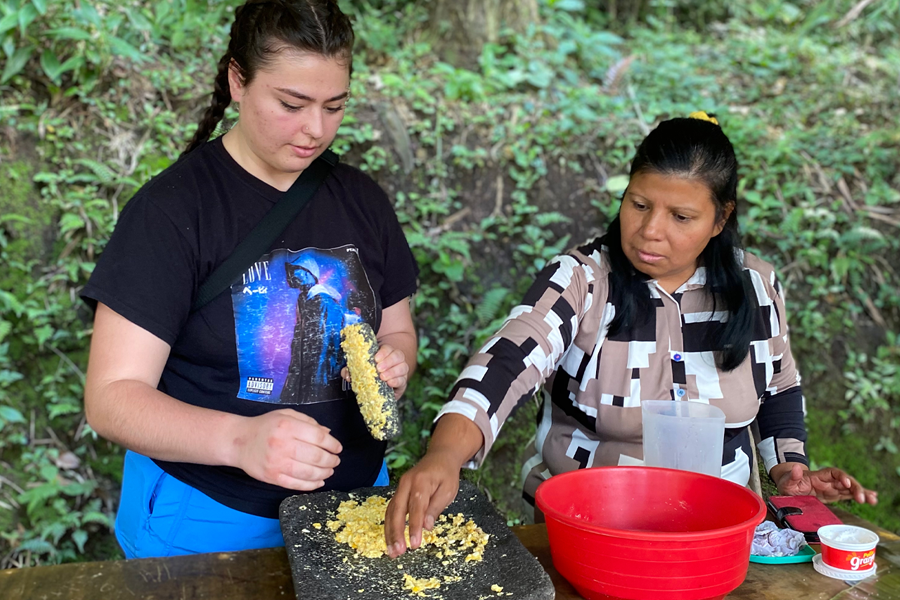 A student and a Native Costa Rican stand outdoors behind a table with a red bowl and ground corn on the table in front of them.
