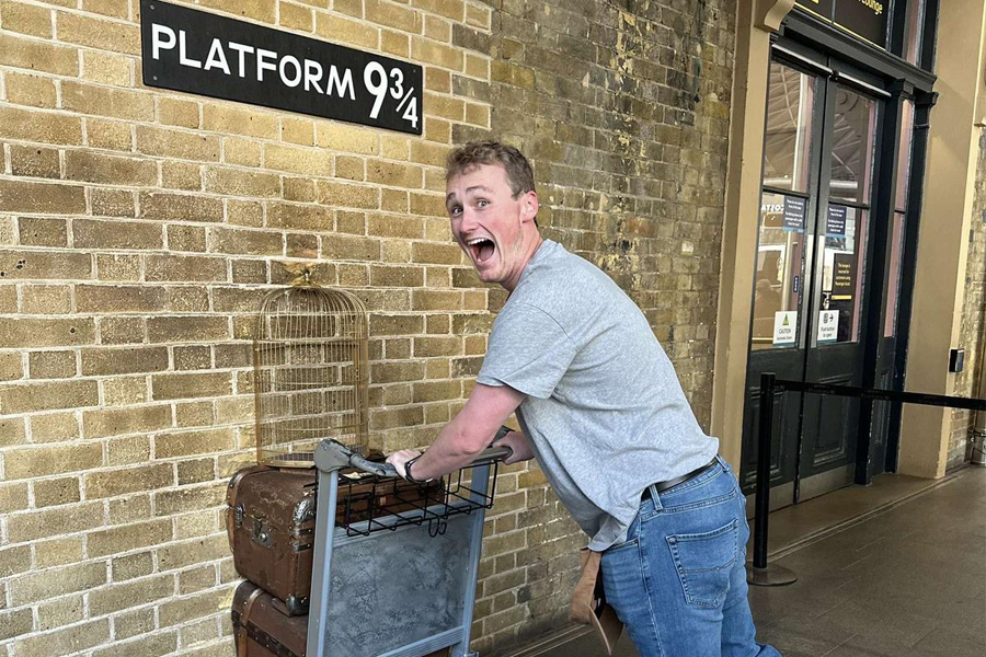 A student pretends to pushes a mock cart that's halfway into a brick wall below a sign that says Platform 9 3/4.