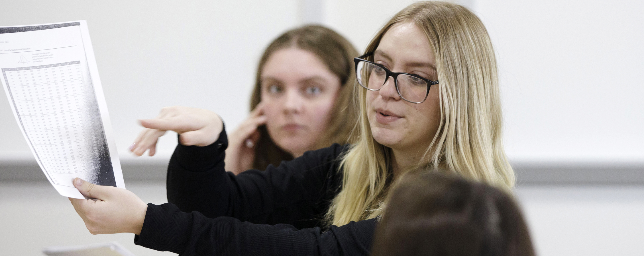 A student wearing glasses holds and points at a piece of paper.