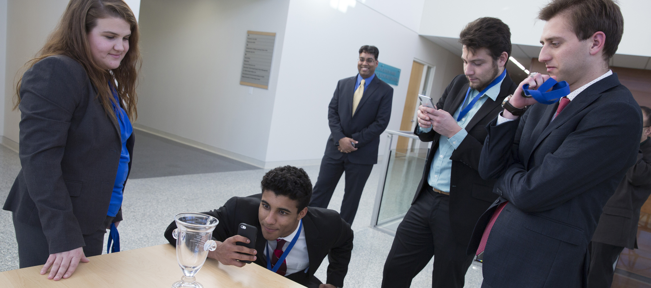 A small group of students gather around a table with a clear trophy with a smiling faculty member in the background.