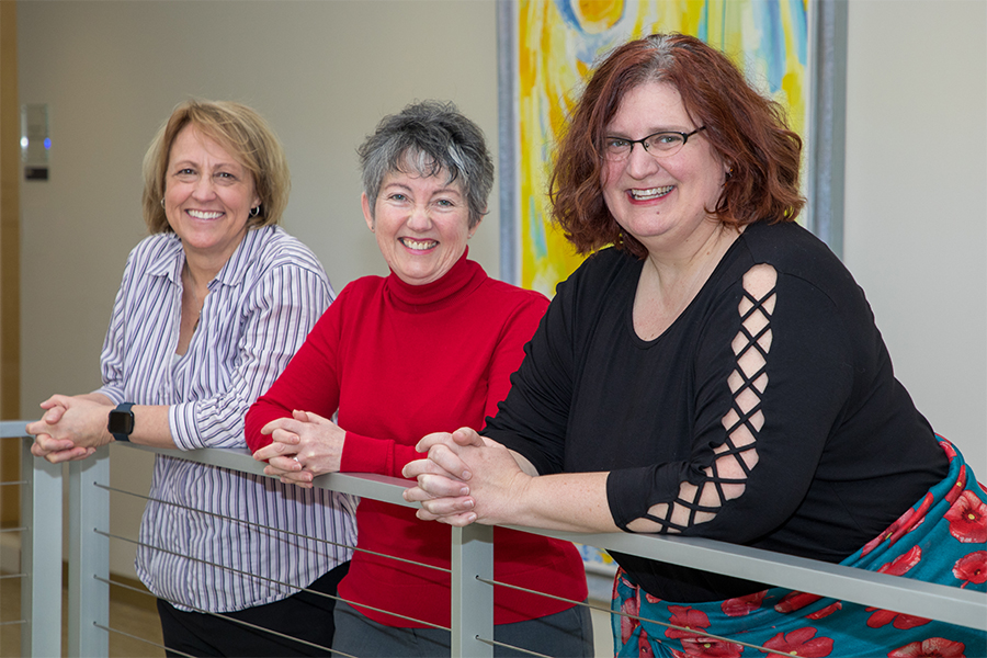 Three people lean over a stair railing and smile at the camera.