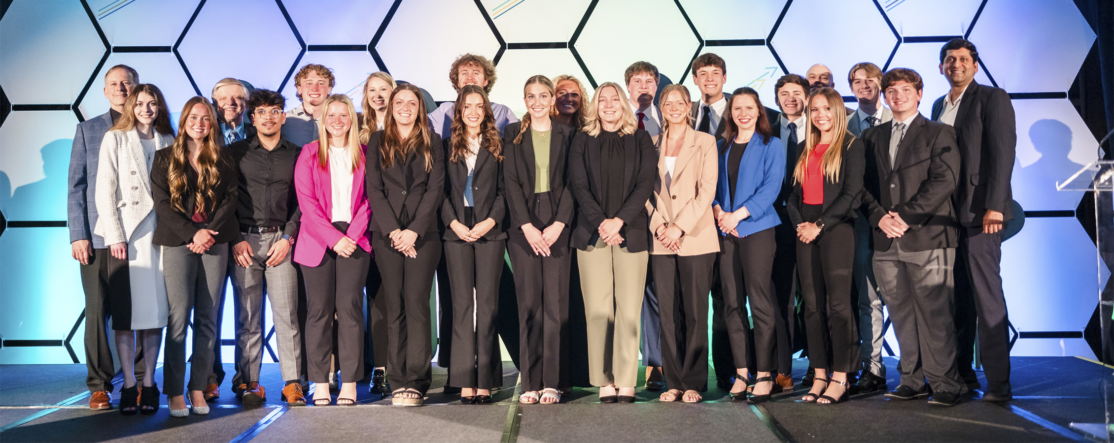 A large group of students stand together facing the camera with a colorful hexagon background.