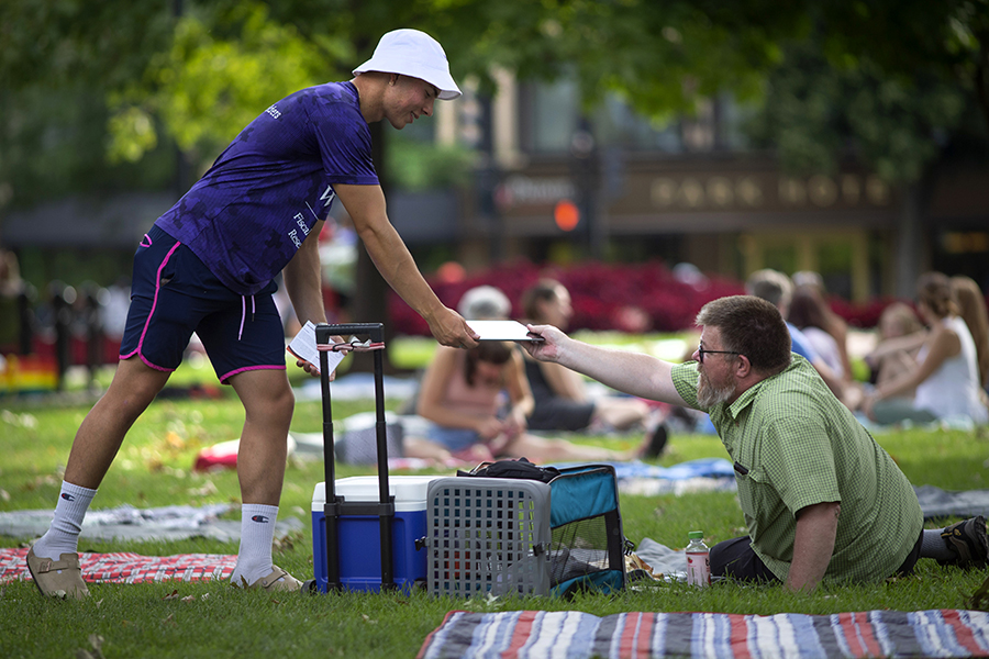 An economics student wearing a purple shirt and white hat hands a survey to someone sitting in a lawn chair at a park.