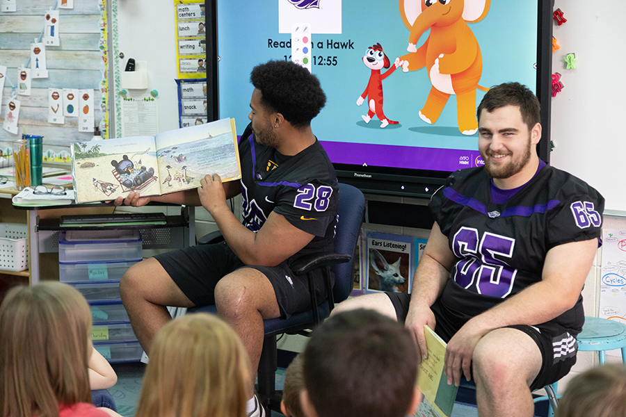 Two UW-Whitewater students wearing Warhawk football jerseys read a book to a classroom of children.