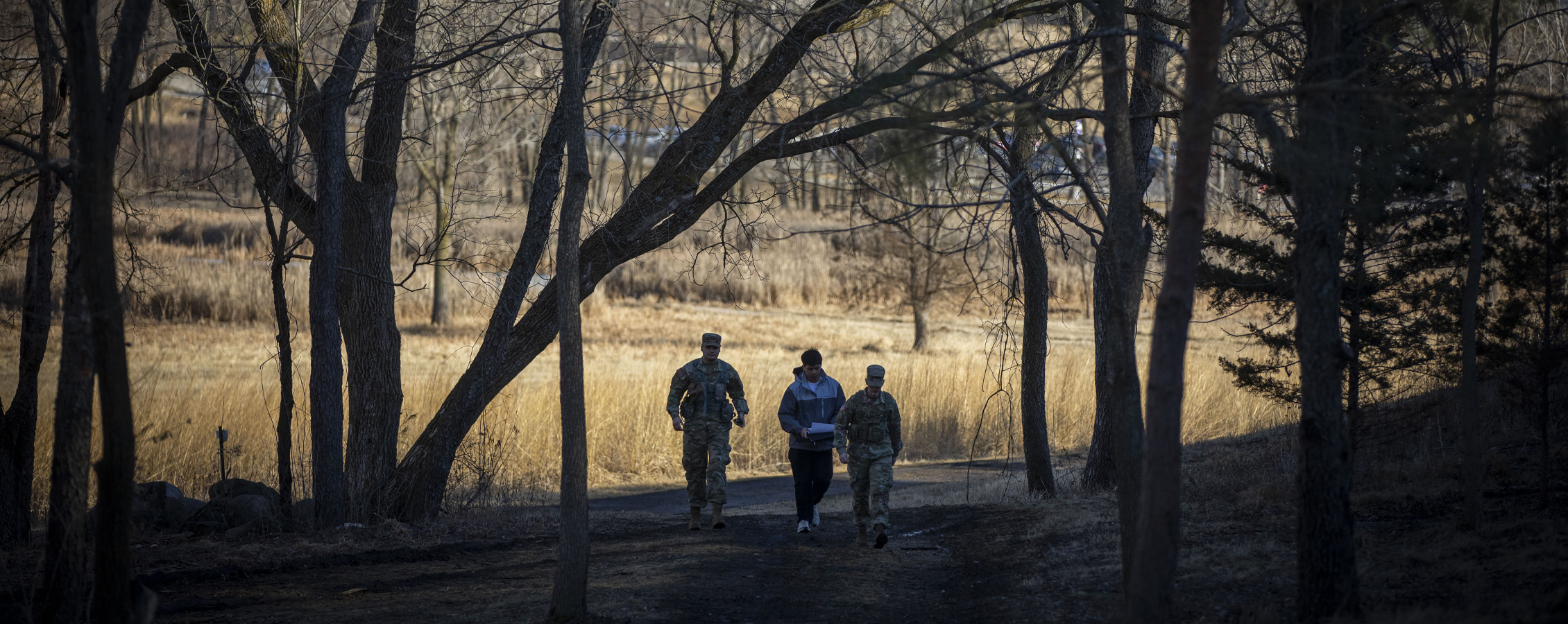 Two people dressed in camo fatigues walk through a path in the woods with another person.