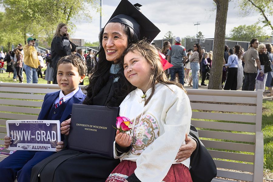 An adult graduate dressed in cap and gown sits on a bench, smiling, with two children.
