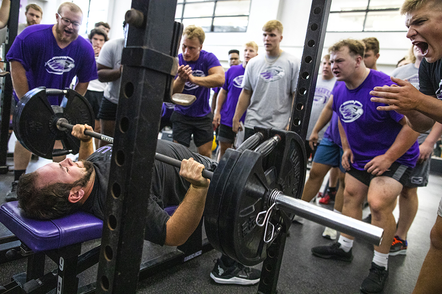 A group of crowd around another student benching a large amount of weight. 