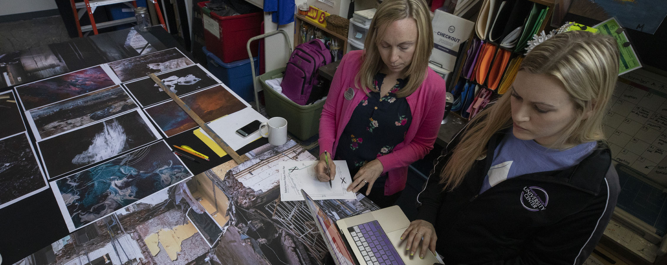 Two people work at a long table with a laptop and pictures laid out.