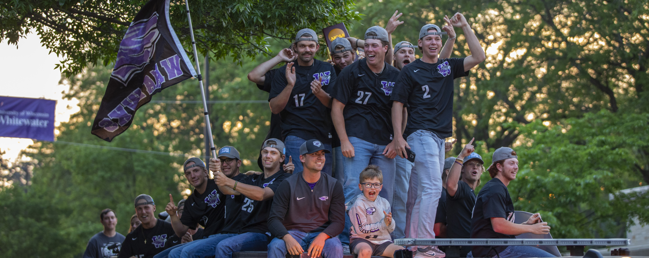 Warhawk Baseball members celebrate while they ride in a truck and hold their NCAA Championship trophy.