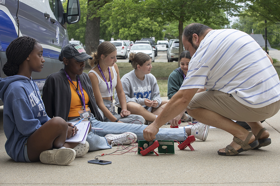 A faculty member kneels on the sidewalk with several precollege attendees, showing them electrical wiring.