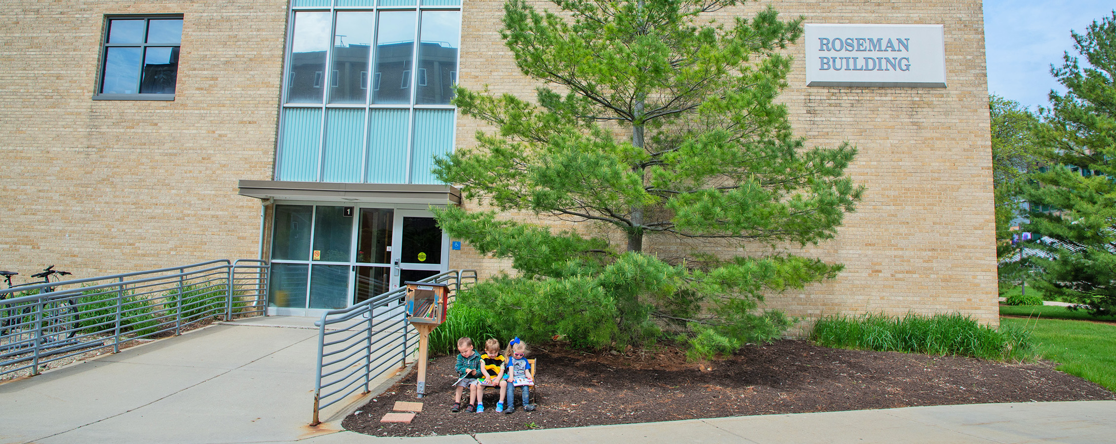 Image outside of Roseman hall with a green tree in front of it and three children sitting in front of the tree.