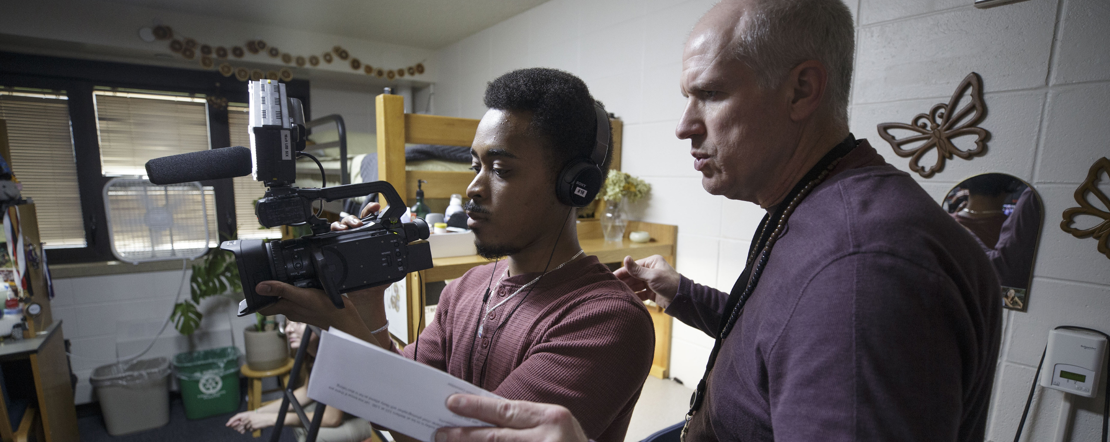 A faculty member stands behind and directs a student with a video camera.