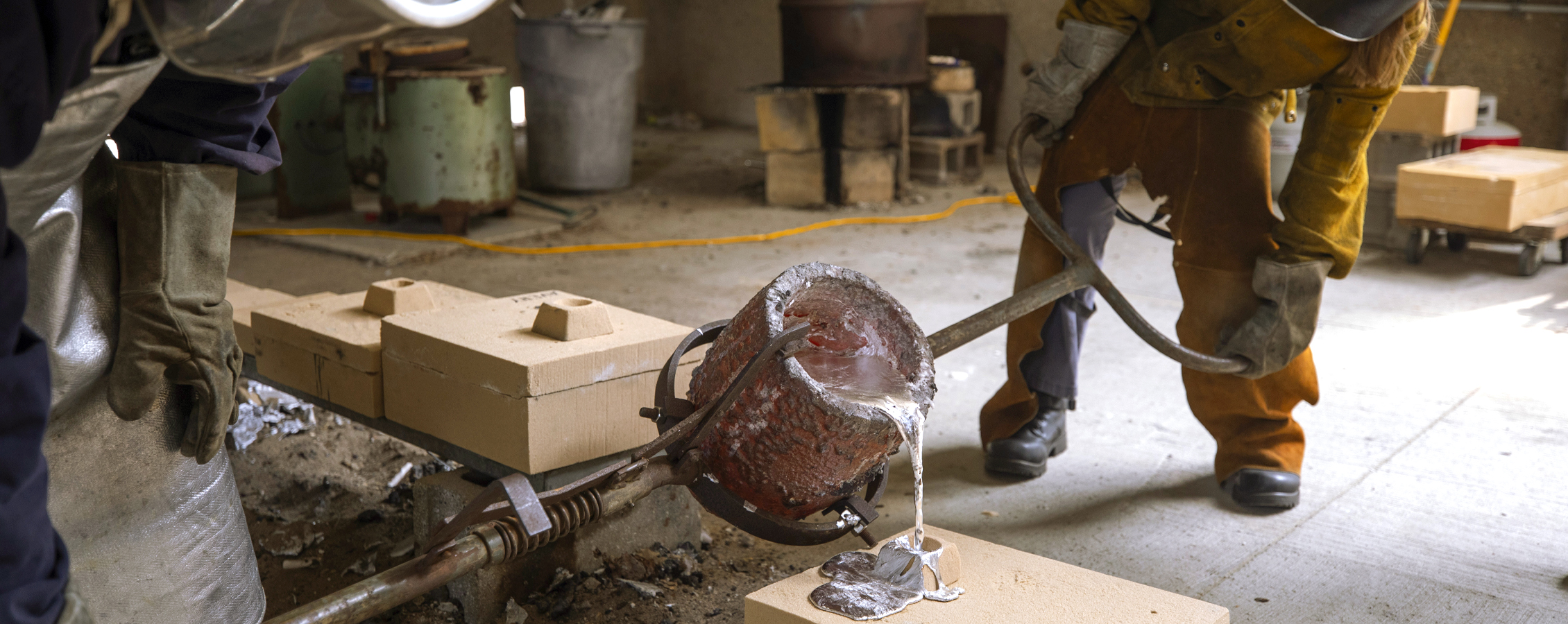 Two people dressed in yellow protective gear use long handles to pour liquid metal.