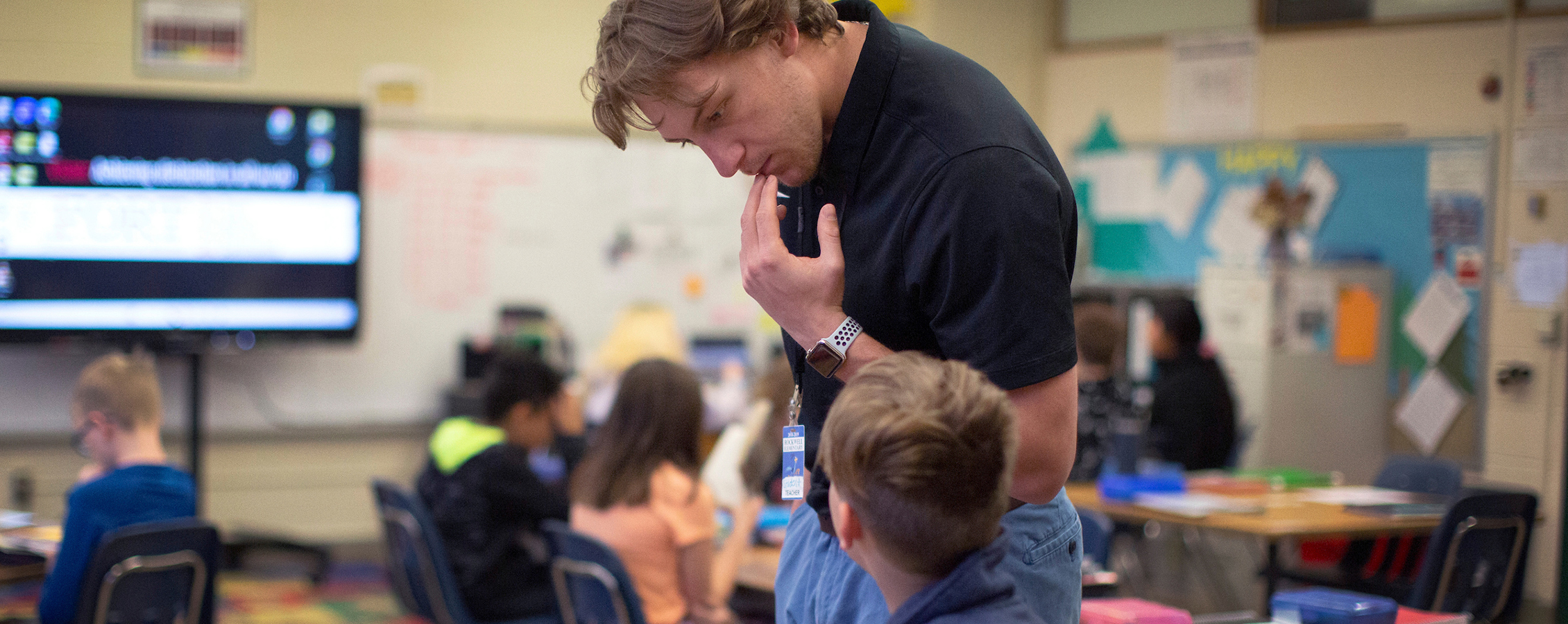 Male teacher in an elementary school classroom looks over the shoulder of a student to check their work.
