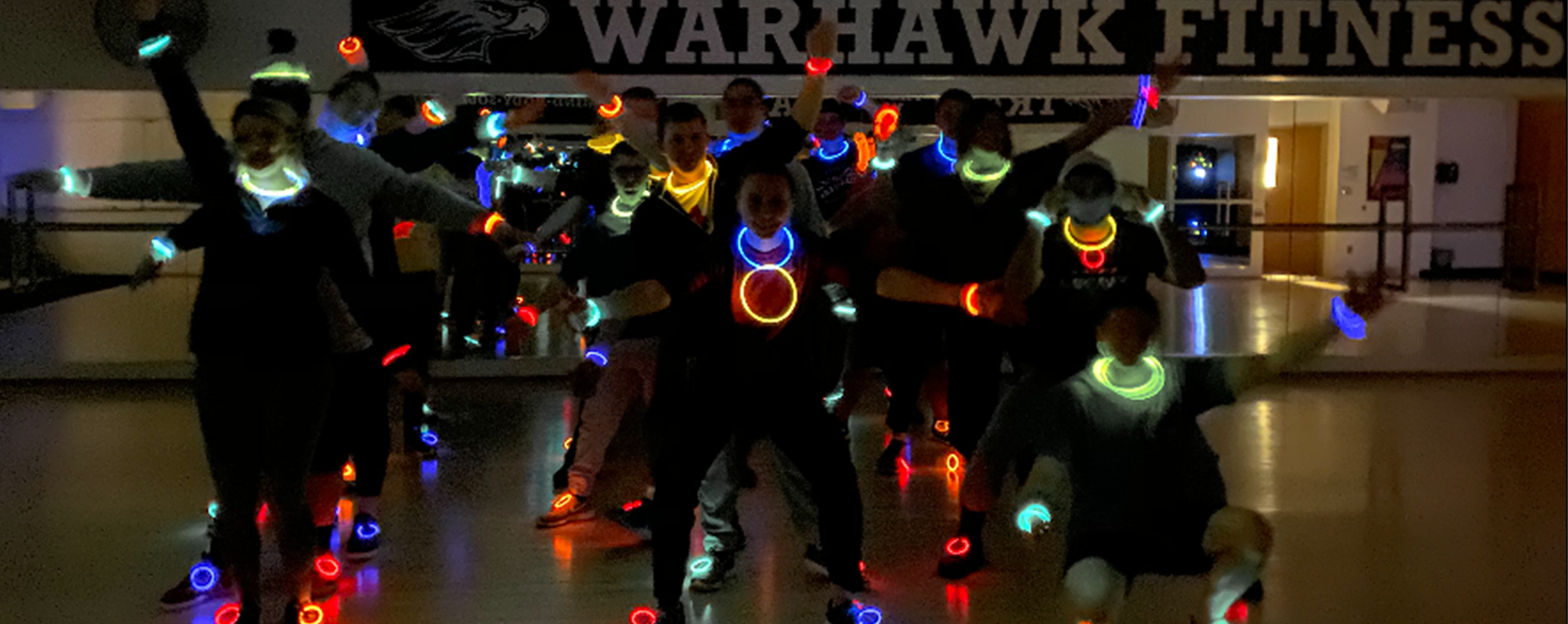 A dark photos of students gathered in front a Warhawk Fitness sign wearing colorful glow sticks.