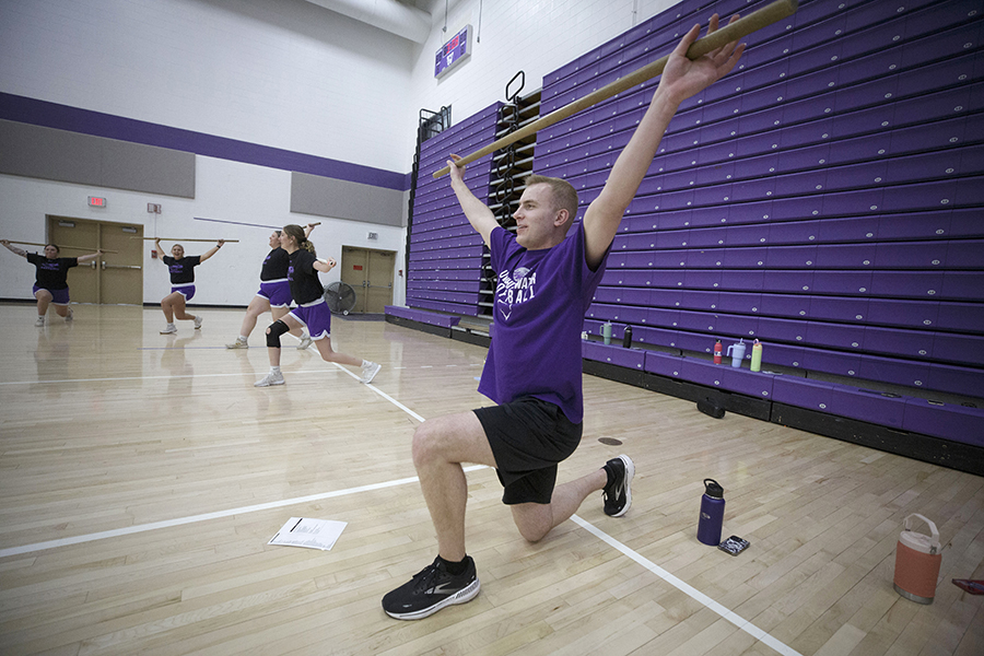 A student in a purple shirt kneels on a gym floor and holds a bar above his head.