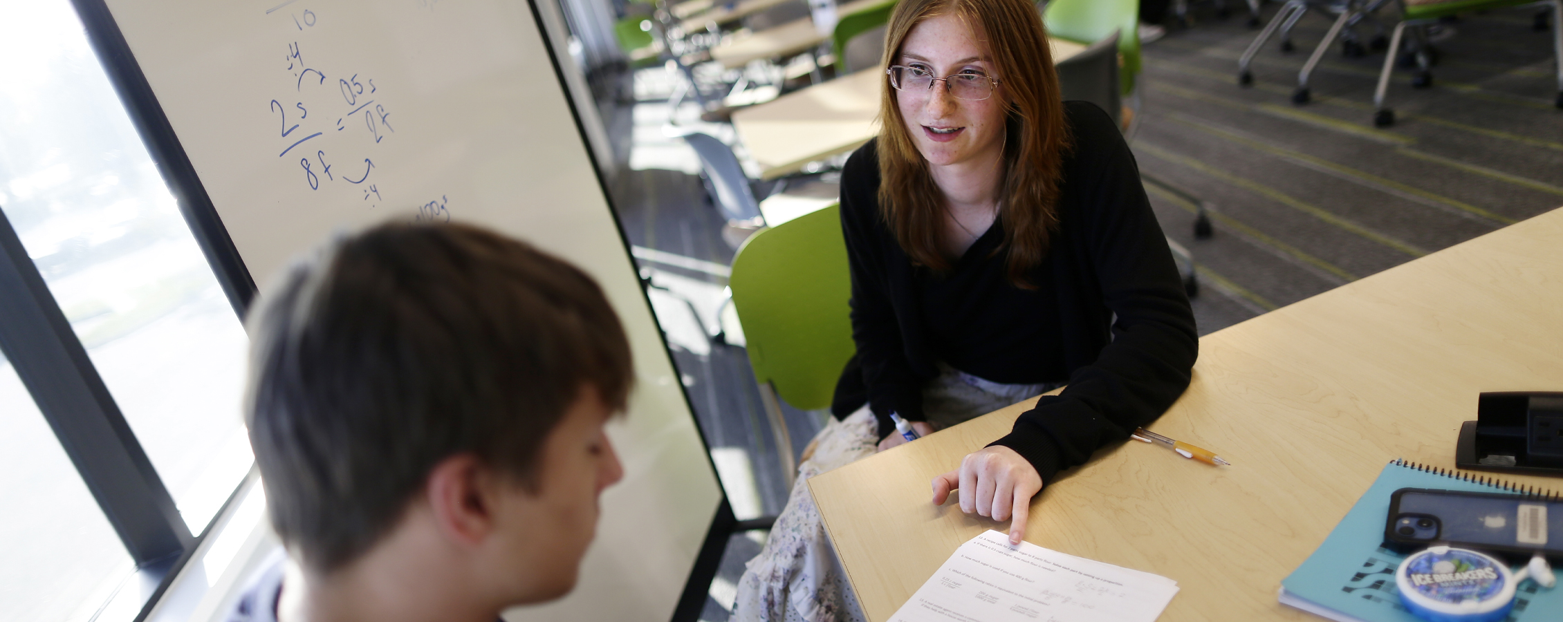 Two people work together at a table with an equation written on a whiteboard behind them.