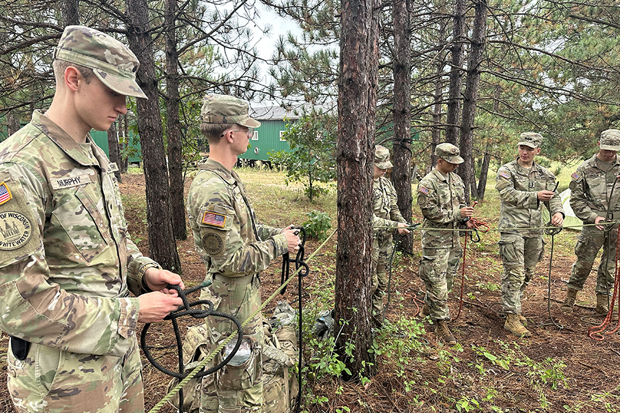 A group of people in camo fatigues stand in a line in the woods while holding rope.