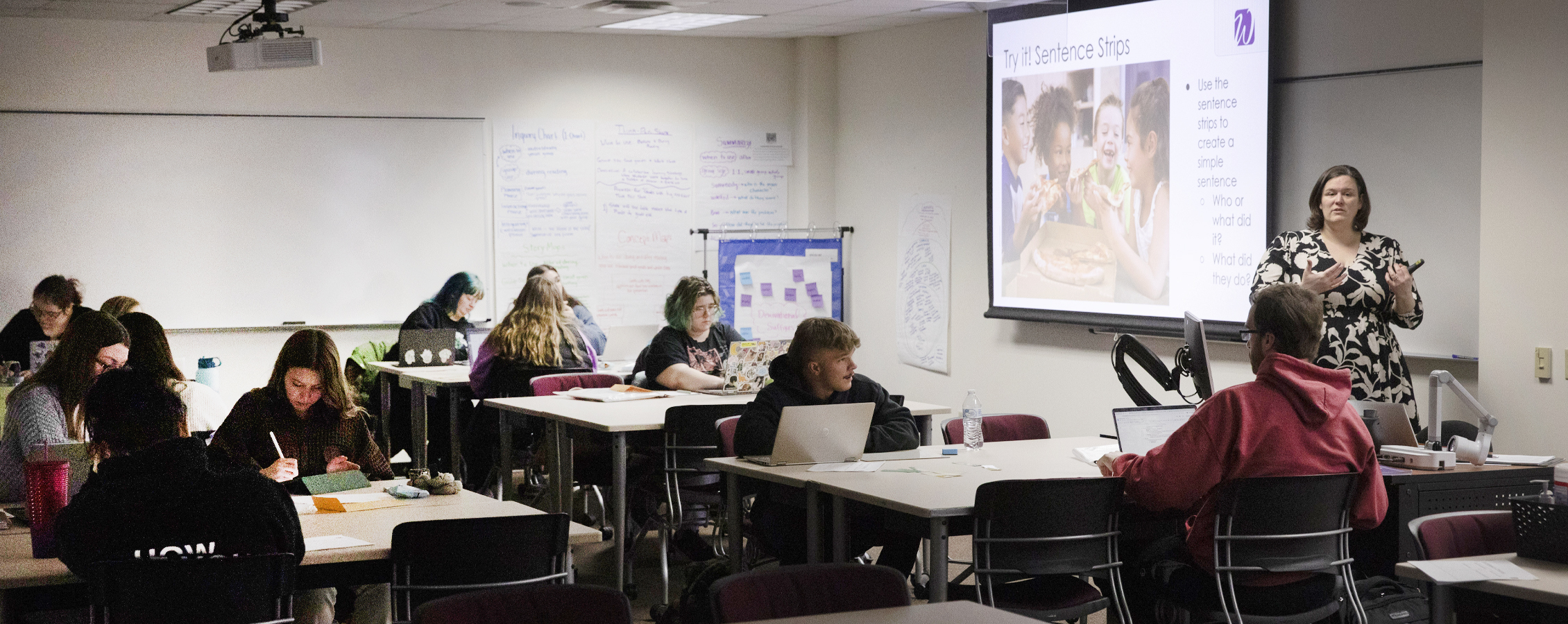 A faculty member speaks at the front of class with her arms spread out.