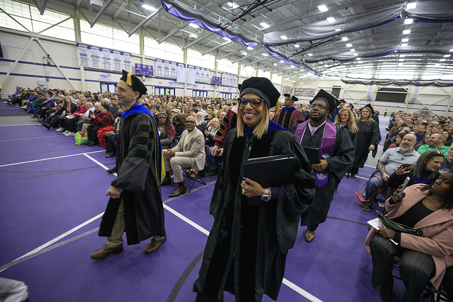 Monica Kelsey-Brown, wearing academic regalia, walks through the fieldhouse during commencement.