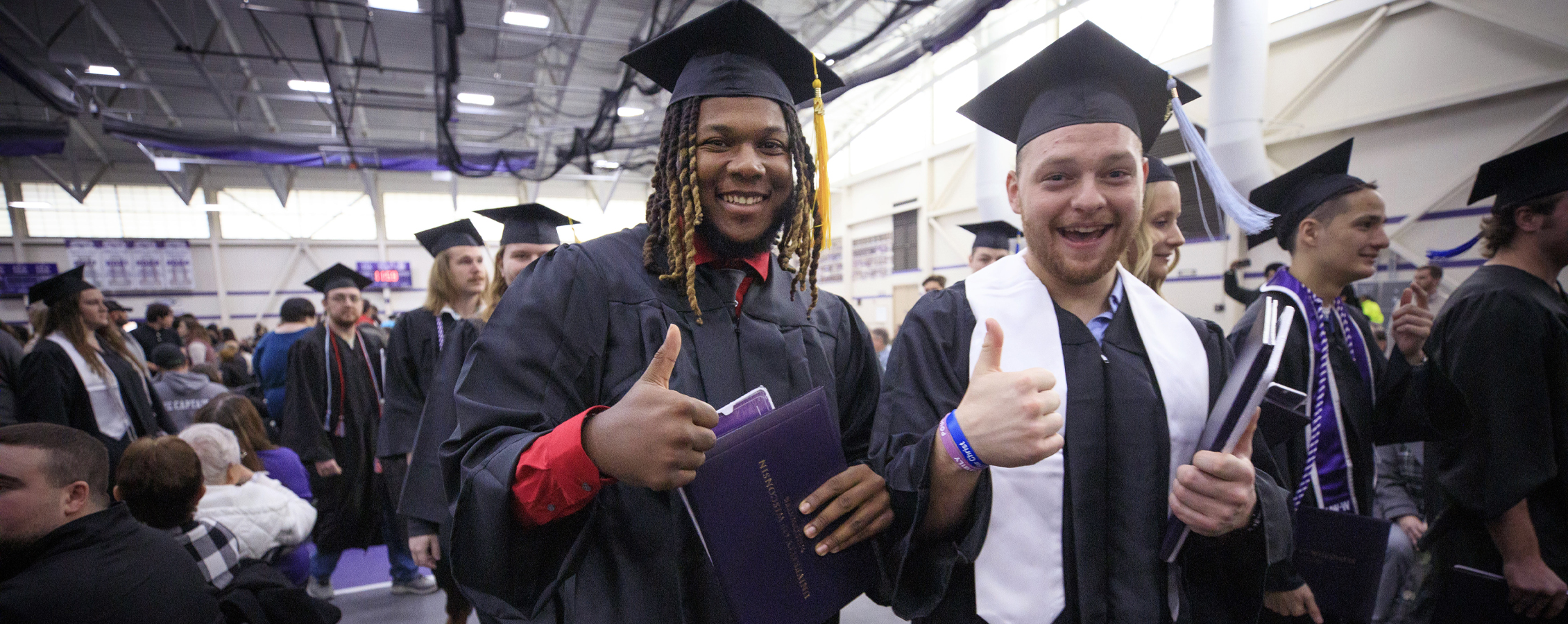 Two graduates, dressed in cap and gown, give a smile and a thumbs up to the camera.