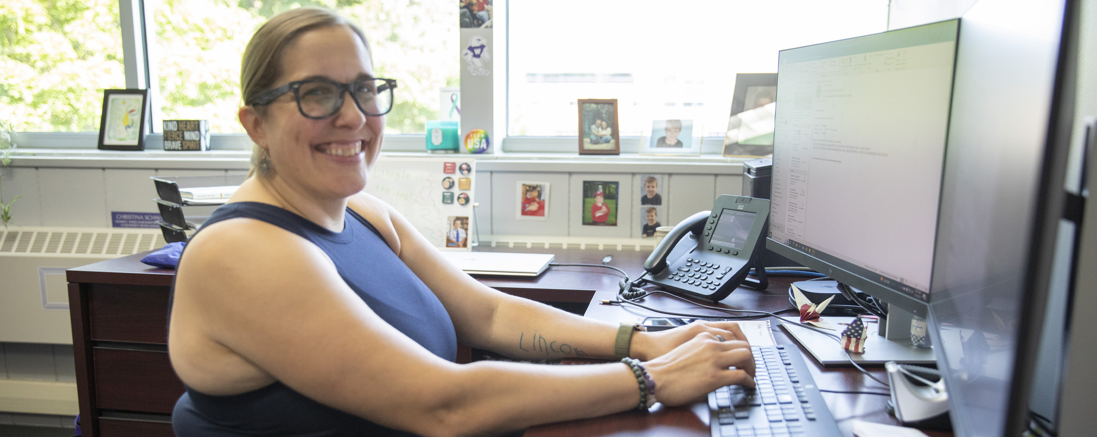 A person sits at desk with their hands on the keyboard and smiles at the camera.