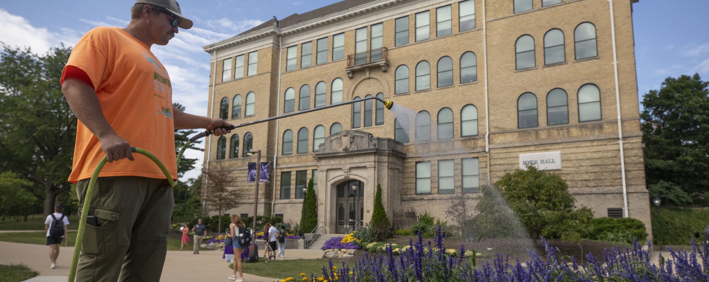 A person waters purple flowers in front of Hyer Hall.