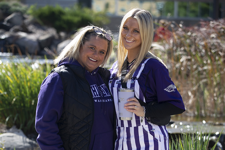 Two women outside at a tailgate at a football game, one wearing a purple sweatshirt and one in purple and white overalls.