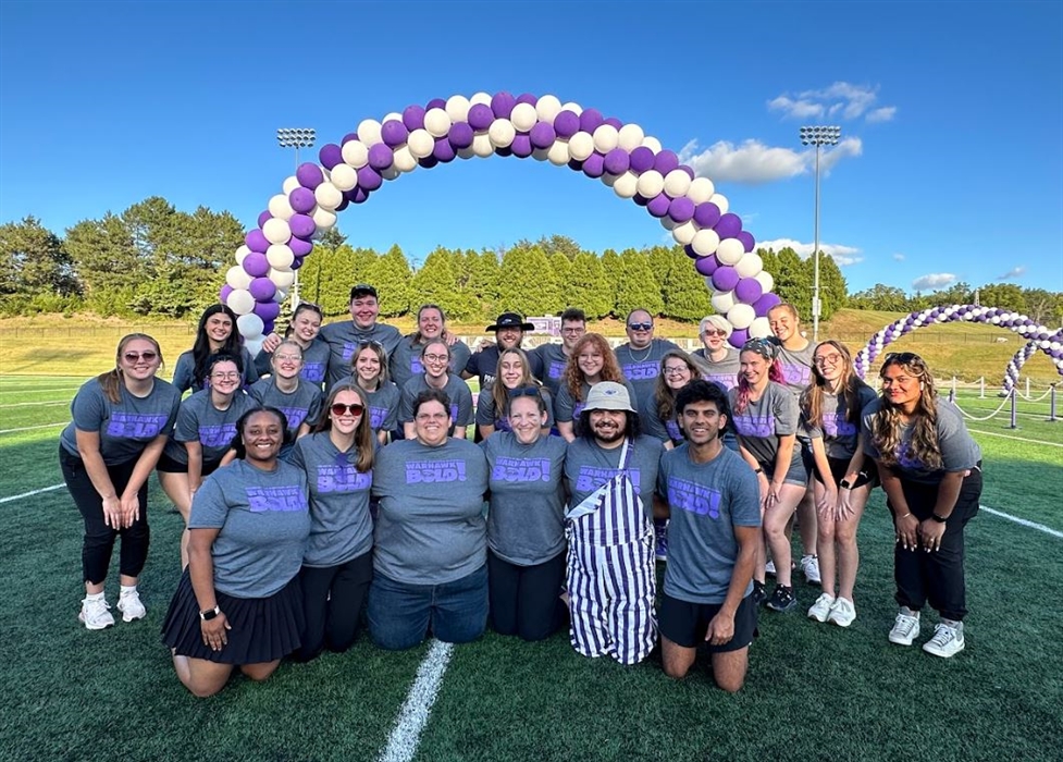 Group of people in purple and gray shirts posed under purple and white balloon arch on football field