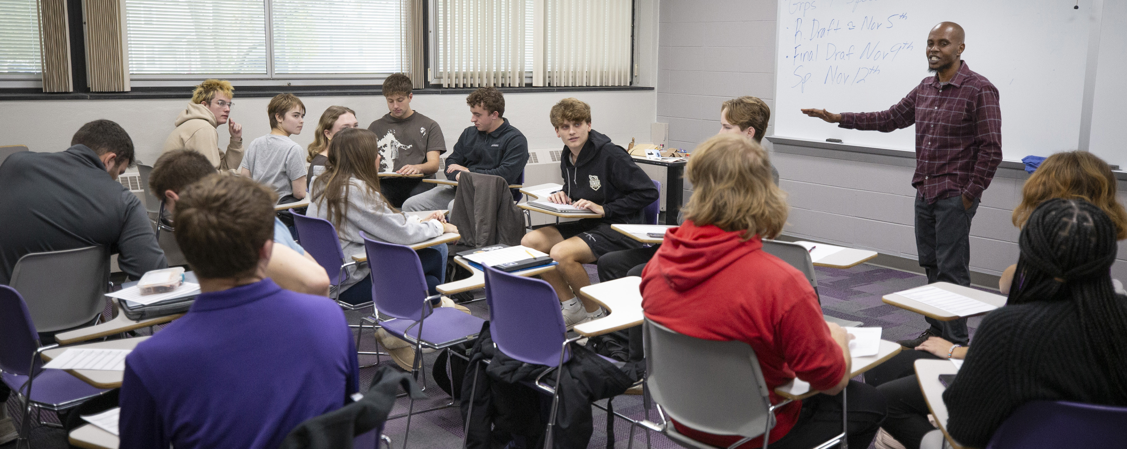 A faculty member teaches in front of a white board while students group together at their desks.