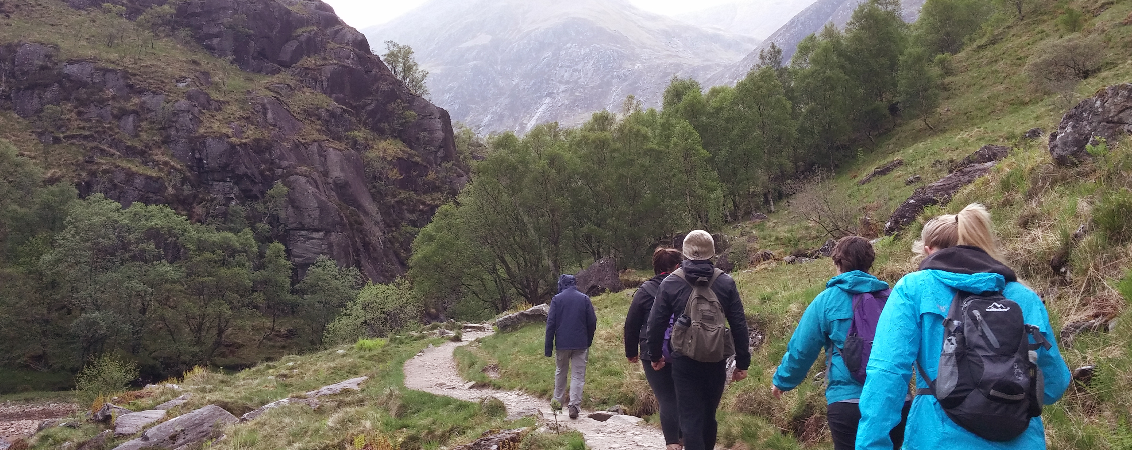 A group of people hike on a worn path amidst mountains and clouds.