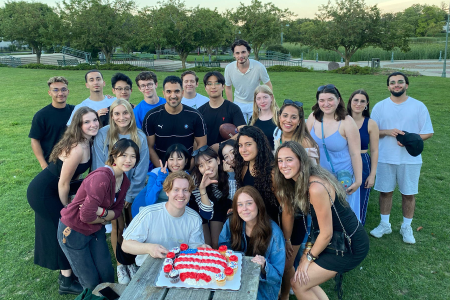 A group of international students celebrate with a red, white, and blue cake in a park.