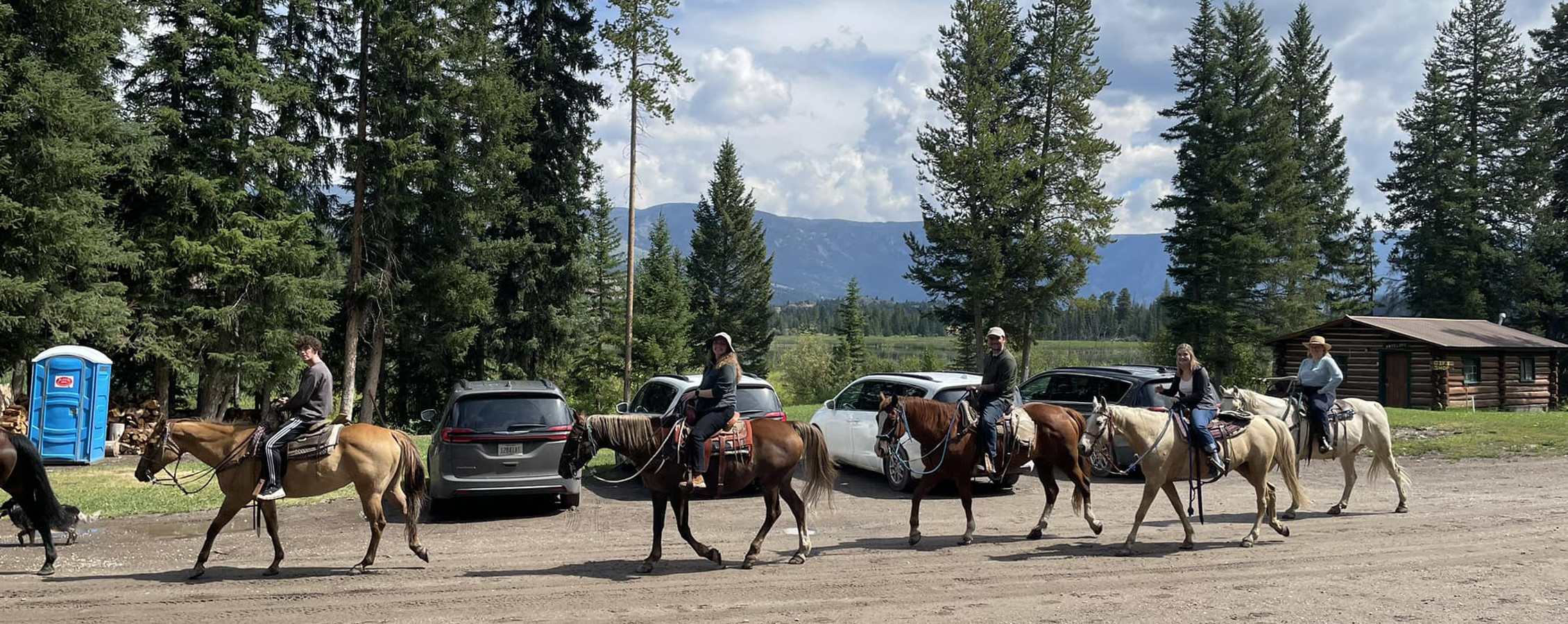A group of people ride horses amidst mountains and evergreen trees.