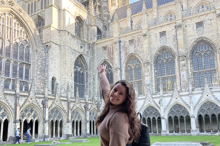 A young woman stands in a courtyard and gestures up toward a European cathedral.