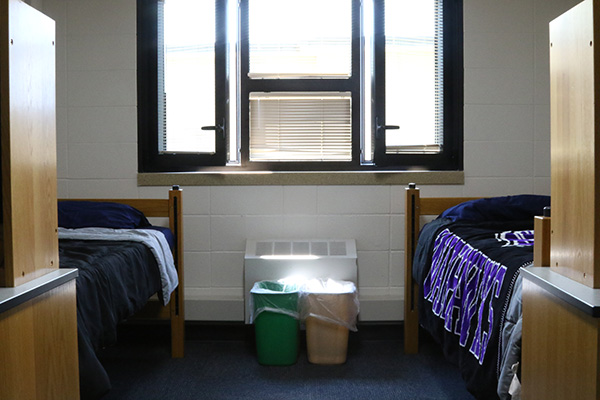 A dorm room with 2 beds and 2 desks, and 2 waste bins in it.