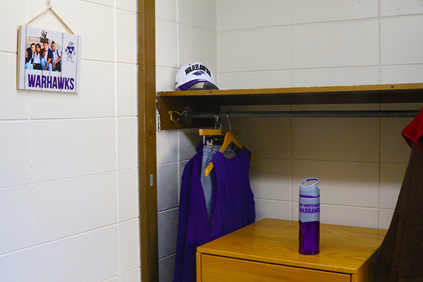 A wells hall dorm closet. There is a self in the closet with a dresser underneath it. There is a water bottle on top of the dresser. The shelf has a bar under it for hanging shirts, and there are multiple shirts hanging in this photo