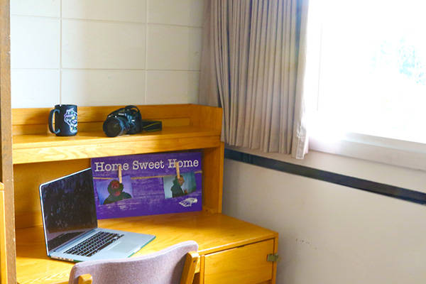 A desk with a laptop and a picture on it. The desk also has a small overhang, where a mug and a camera are sitting.
